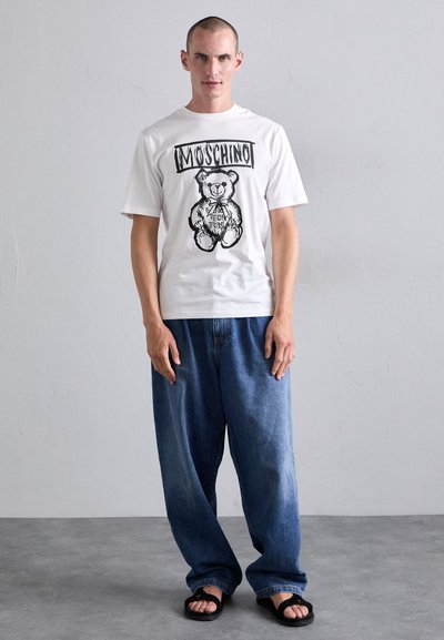 Cămașă albă cu un grafic de urs negru și textul "Moschino", purtată cu blugi lejeri din denim albastru și șlapi negri. Textură netedă.