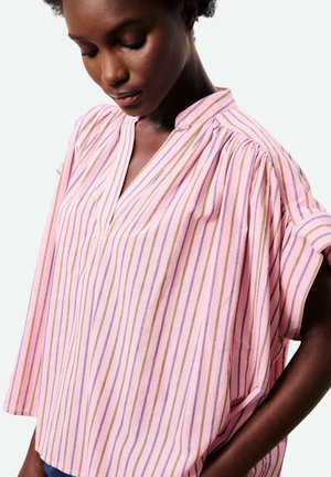 Vrouw in een losse roze blouse met verticale blauwe en gouden strepen, die omlaag kijkt met haar hand in haar zak tegen een witte achtergrond.