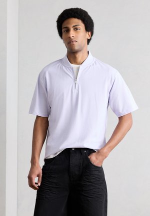 Mężczyzna o afro fryzurze, ubrany w jasnofioletową koszulę z krótkim rękawem na zamek i czarne spodnie, stojący z jedną ręką w kieszeni.