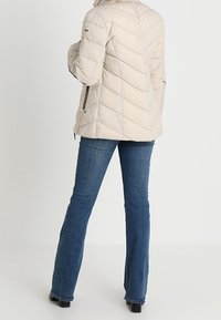 Jasno-beżowa kurtka pikowana w wzór w jodełkę, z bocznymi zamkami i wysokim kołnierzem. Noszona z niebieskimi spodniami dzwonami.