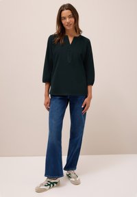 Tunic verde scuro a maniche lunghe con scollo a V e taglio dritto, abbinata a jeans a zampa d'elefante blu e sneakers verdi e grigie. Texture liscia.