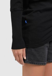 Pull noir avec poignets côtelés, associé à un short en denim foncé. Comprend une petite étiquette bleue sur le côté, une texture lisse et une coupe décontractée.