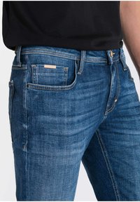 Vaqueros de mezclilla azul oscuro con un corte ajustado. Las características incluyen bolsillos delanteros y traseros, costuras en contraste y una pequeña etiqueta de marca en el bolsillo trasero.