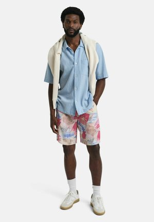 Mann trägt hellblaues kurzärmeliges Hemd mit Knöpfen, mehrfarbige gemusterte Shorts, weiße Sneaker und einen cremefarbenen Pullover, über die Schultern gelegt.