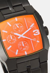 Orologio in metallo nero con cassa quadrata, quadrante arancione vivace, indicatori delle ore bianchi, tre sottomisure e una finestra per la data. Cinturino in gomma testurizzata.