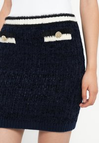 Gonna texturizzata blu scuro con cintura a strisce bianche e nere, tasche decorative con bottoni dorati, indossata con una maglietta bianca.