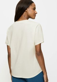 Kremowa koszula z krótkim rękawem, gładka tkanina, okrągły dekolt, detal szwu na plecach, lekko luźny krój, zestawiona z niebieskimi spodniami.