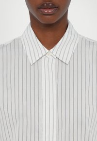 Gros plan d'une personne portant une chemise blanche boutonnée avec de fines rayures verticales noires et un col pointu.