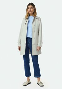 Hellgrauer Trenchcoat mit durchgehender Knopfleiste, schlankem Schnitt und klassischem Kragen, kombiniert mit einem hellblauen Oberteil und dunklen Jeans.