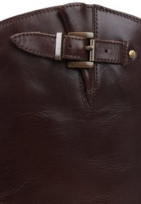 Bota de couro marrom com uma superfície texturizada, apresentando um detalhe de fivela e tira metálica na parte superior. Bordas costuradas são visíveis.