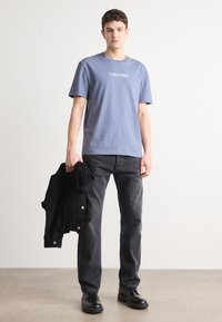 Gaiši zila kokvilnas T-kreklu ar baltu "Calvin Klein" logo, apvienots ar melnām džinsa biksēm un melnu jaku, valkāts ar melnām kurpēm.