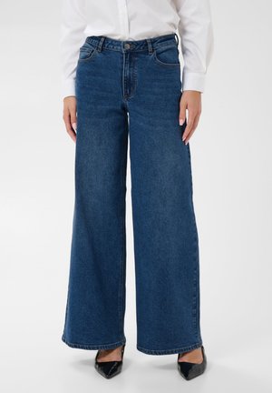 Jean bleu à jambes larges porté avec une chemise blanche boutonnée et des chaussures noires pointues, montré de la taille jusqu’aux pieds.