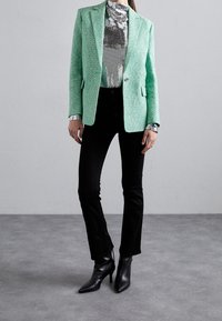 Mátově zelený blazer s texturovanou tkaninou, doplněný o stříbrný top s flitry a černé rozšířené kalhoty. Černé špičaté kotníkové boty celý vzhled dotvářejí.