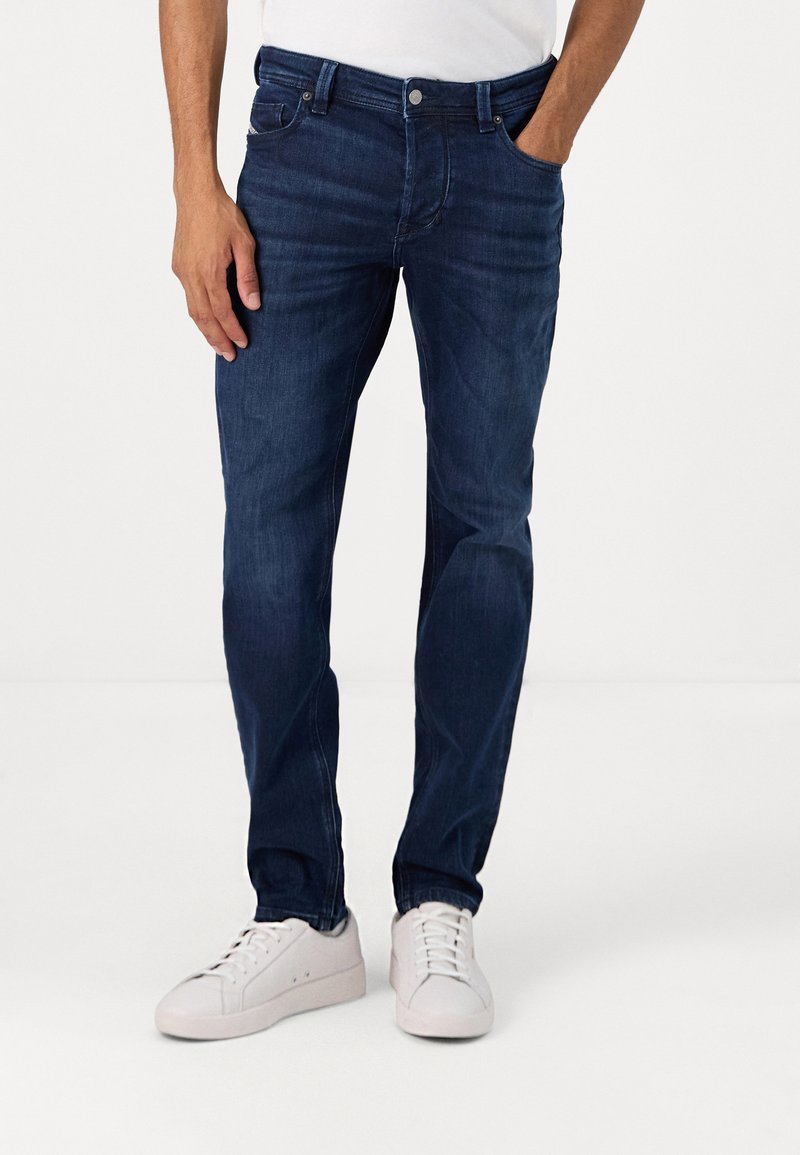 Jeans de mezclilla azul oscuro con piernas rectas, diseño de cinco bolsillos y cierre clásico de botón y cremallera. Usados con zapatillas blancas.