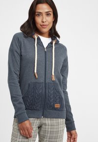 Veste à zip marine avec des poches en tricot �à motif torsadé, des cordons de serrage en rope contrastants et un patch en cuir au niveau de la poche inférieure.