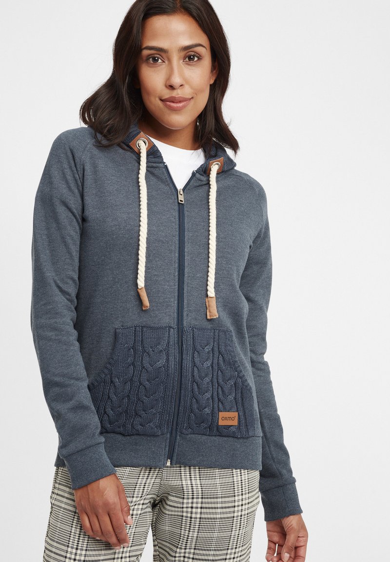Veste à zip marine avec des poches en tricot à motif torsadé, des cordons de serrage en rope contrastants et un patch en cuir au niveau de la poche inférieure.