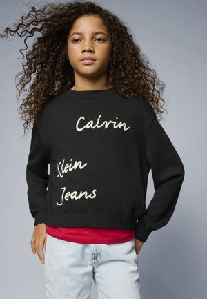 Black knitted sweater featuring white embroidered text: "Calvin Klein Jeans." Worn over a red shirt, paired with light blue jeans.