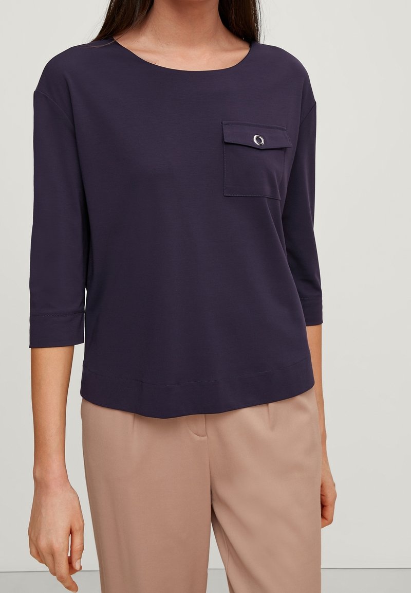 Femme portant un haut bleu marine foncé à manches trois-quarts avec une poche poitrine et un pantalon beige clair sur un fond uni.