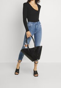 Bolso tote negro con estampado de cocodrilo, con dos asas resistentes, forma triangular y acabado brillante. Lleva puesto con jeans azules rasgados y zapatos de tacón negros abiertos.