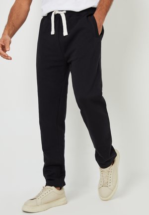 Man draagt zwarte joggingbroek met witte trekkoord en beige sneakers, loopt met één hand in de zak tegen een grijze achtergrond.