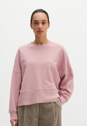 Vrouw met donker haar naar achteren gebonden, draagt een losse roze trui en beige ribfluwelen broek tegen een effen witte achtergrond.