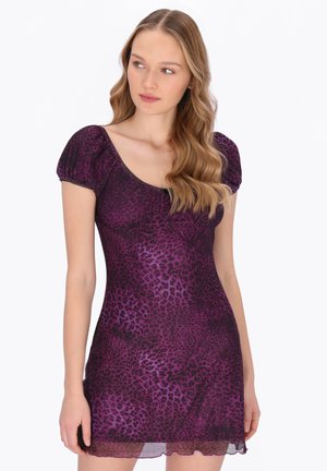 Ung kvinde med langt, bølget hår iført en kort, lilla kjole med leopardprint og puffede ærmer, der kigger til siden.