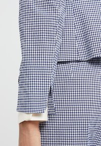 Námořnický a bílý kostkovaný blazer s přiléhavým střihem. Má záhyb na rukávu a pod ním je vidět bílý manžet. Těžká strukturovaná tkanina.