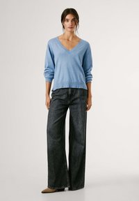 Mėlynas mezginys su V formos kaklu ir nuleistais pečiais, derinamas su aukštu liemeniu, plačiomis tamsiomis džinsomis ir smailia forma rudos spalvos bateliais.