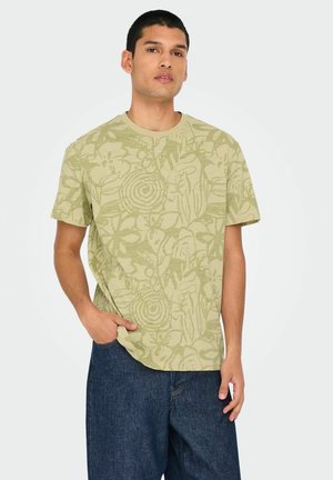 Camiseta verde claro con un patrón botánico, cuello redondo, mangas cortas y corte relajado. Combinada con pantalones cortos de mezclilla oscura.