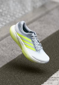 Svetlo siva športna obutev z neonsko rumenimi poudarki, elegantno zasnovo, zgornji del iz zračne mrežice in teksturiran podplat. Na voljo z ikoničnimi tremi črtami.
