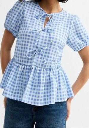 Femme portant un chemisier à manches courtes à carreaux bleu et blanc avec trois liens devant et une taille à basque, assorti à un jean bleu foncé.
