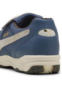 Sapatilha azul em couro com uma faixa em camurça com detalhes dourados e fecho de velcro. Apresenta uma sola arredondada e acolchoada, além de um logo na parte traseira.