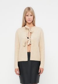 Bežs adīts džemperis ar auklas aizdari, kurā redzama teksturēta raksts. To valkā virs melnas piegulošas svārku, ar taisnu piegriezumu un atvērtu priekšpusi.