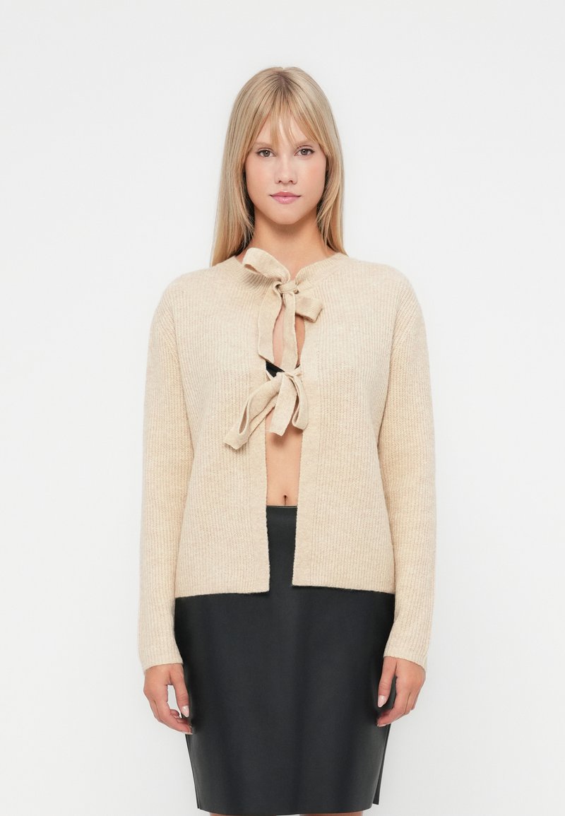 Bežs adīts džemperis ar auklas aizdari, kurā redzama teksturēta raksts. To valkā virs melnas piegulošas svārku, ar taisnu piegriezumu un atvērtu priekšpusi.