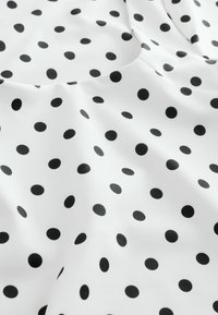 Tissu blanc à pois noirs uniformément espacés, présenté avec des plis doux et une texture lisse.