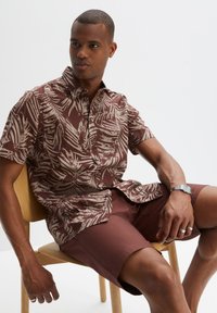 Camisetta marrone a maniche corte con motivo di foglie di palma bianche, abbinata a pantaloni corti marroni coordinati. Una sedia in tonalità legno naturale esalta lo stile minimalista.