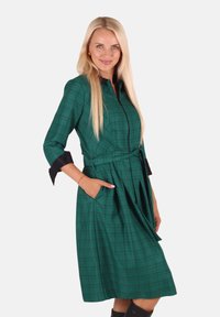 Grünes kariertes Kleid mit langen Ärmeln, schwarzen Akzenten, tailliertem Bund und seitlichen Taschen. Verfügt über ein schürzenartiges Rockdesign.