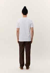 Persona stāv ar muguru pretējā virzienā, tērpusies baltā T-kreklā, tumši brūnās biksēs, melnā cepurē un brūnās kurpēs uz gaiša, vienkārša fona.