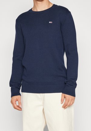 Homme portant un pull bleu marine à col rond avec un petit logo sur la poitrine et un pantalon blanc, debout devant un fond uni.