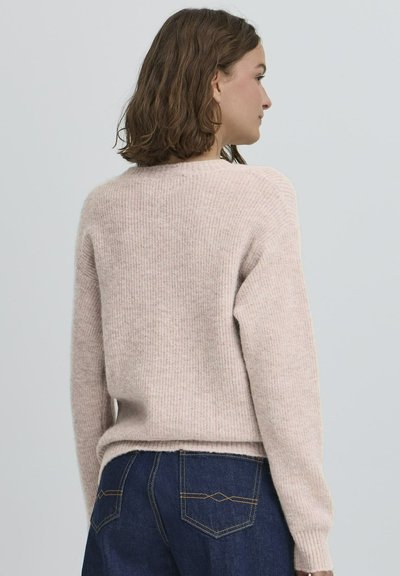 Moteris su pečius siekiančiais rudais plaukais, dėvinti šviesiai rožinį megztinį ir tamsiai mėlynas džinsus, matoma iš nugaros prieš paprastą foną.