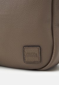 Borsa in pelle marrone con una finitura testurizzata, dotata di un'etichetta quadrata con la scritta "FREDs BRUDER" in lettere stampate in rilievo. Angoli arrotondati, design semplice.