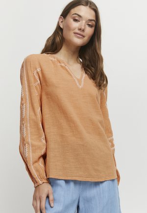 Orange linned langærmet skjorte med V-hals, broderede hvide mønstre på ærmerne og en afslappet pasform. Sammen med lyseblå bukser.