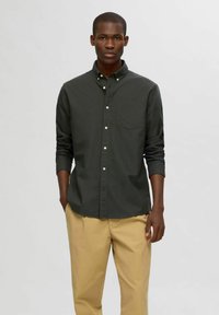 Camisa de botón verde oscuro con manga larga y un bolsillo en el pecho, combinada con pantalones beiges. Textura suave, corte clásico y diseño simple.