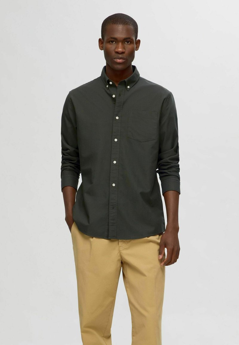 Camisa de botón verde oscuro con manga larga y un bolsillo en el pecho, combinada con pantalones beiges. Textura suave, corte clásico y diseño simple.