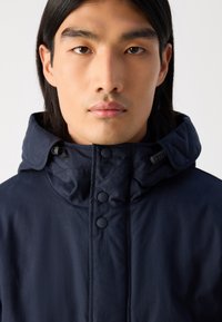 Navy blue jacket with a high collar, buttoned front, and adjustable hood. Smooth fabric with minimal seams and no visible patterns or accents.