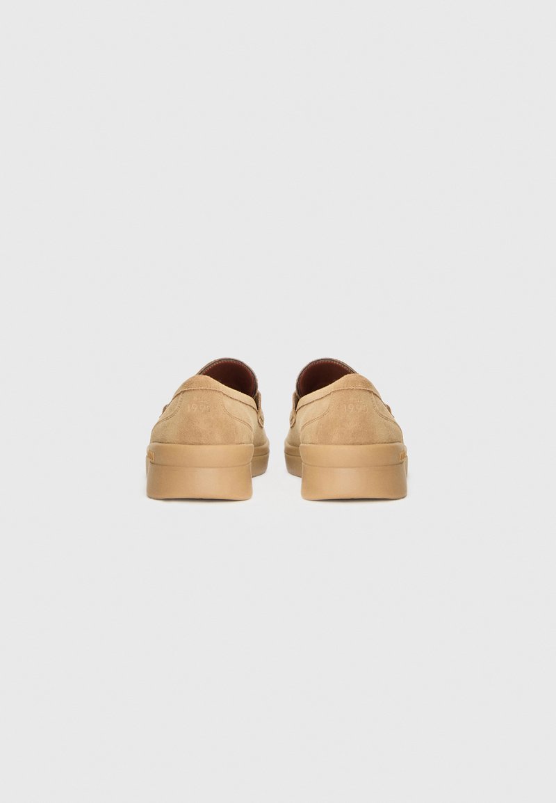 Zapatos slip-on de ante color beige con forma redondeada, que cuentan con una suela de goma beige y detalles en relieve en el talón. El interior es de color marrón oscuro.