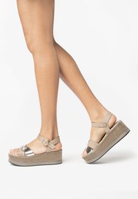 Béžové platformové sandály s kovovým páskem na prstech a nastavitelným řemínkem kolem kotníku. Měkký semišový povrch s robustní podrážkou, která má šedý základ.