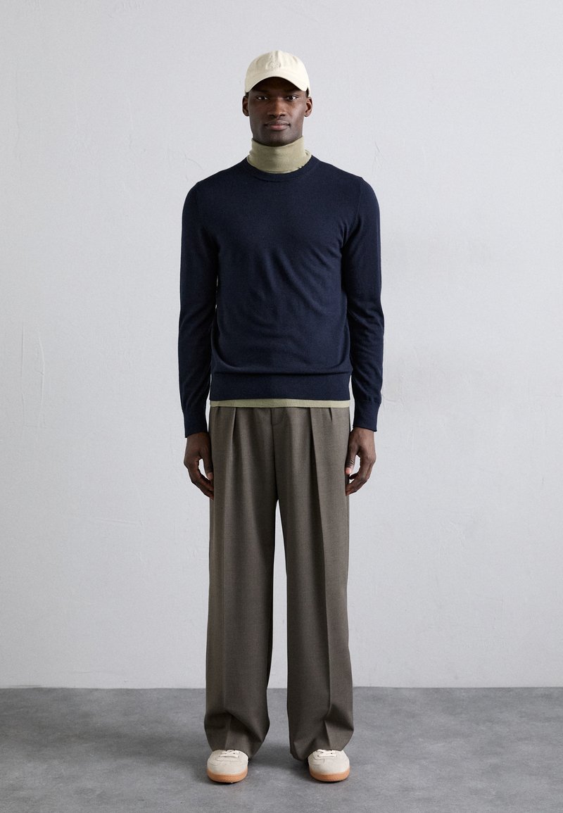 Jersey de cuello redondo en color navy superpuesto a un suéter ligero de cuello alto, combinado con pantalones anchos marrones y deportivas crema, rematado con una gorra beige.