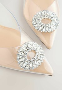 Priehľadné béžové topánky so špicatou špičkou, priehľadnými stranami a veľkými okrúhlymi brošňami z brúsených kamienkov na prednej strane.