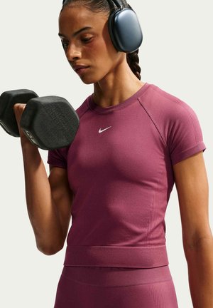 Kobieta nosząca bezprzewodowe słuchawki i fioletowy strój treningowy Nike podnosi czarną hantlę z skupionym wyrazem twarzy.